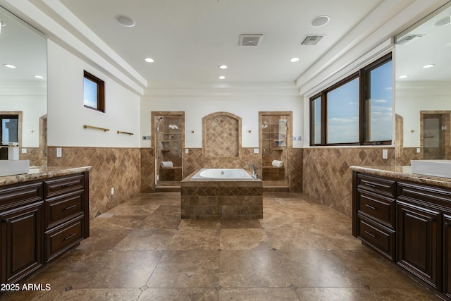 full bathroom with tile walls, two vanities, visible vents, a bath, and walk in shower