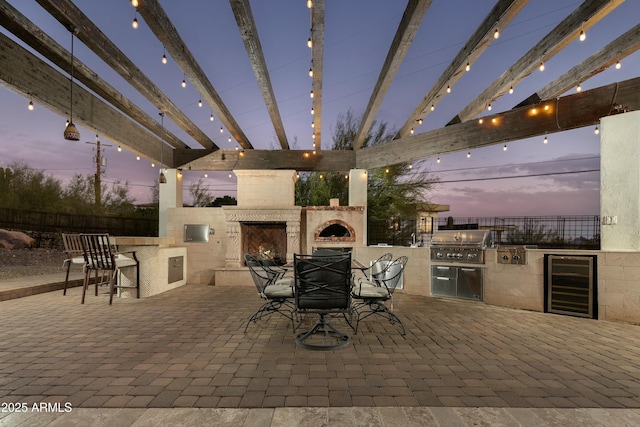 patio terrace at dusk featuring wine cooler, fence, area for grilling, and an outdoor fireplace