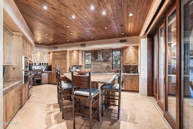 kitchen with wall chimney exhaust hood, a center island, light stone counters, tasteful backsplash, and a breakfast bar area