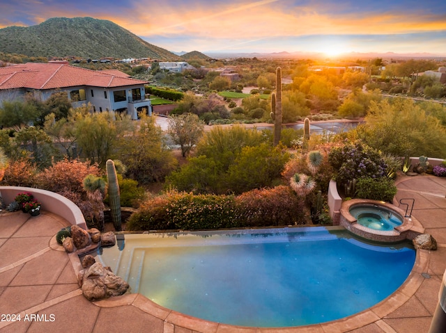 pool at dusk with a water slide, a mountain view, and an in ground hot tub