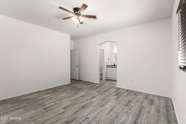 unfurnished bedroom with wood finished floors, baseboards, arched walkways, ceiling fan, and ensuite bathroom