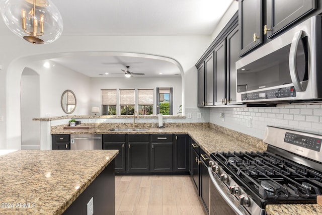 kitchen with backsplash, light stone counters, ceiling fan with notable chandelier, stainless steel appliances, and sink