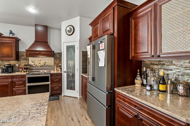 kitchen featuring custom exhaust hood, light hardwood / wood-style flooring, tasteful backsplash, light stone counters, and stainless steel appliances