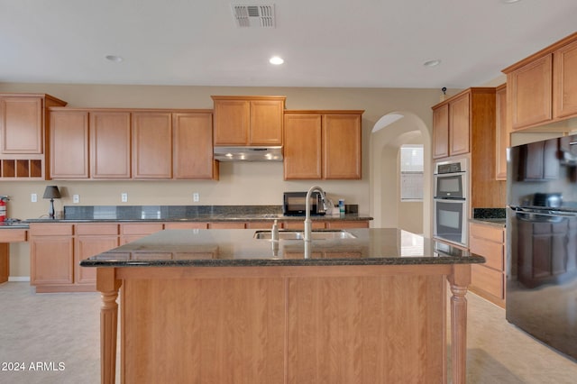 kitchen with double oven, an island with sink, sink, dark stone countertops, and black fridge