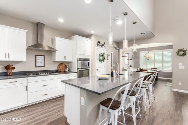 kitchen featuring decorative light fixtures, white cabinetry, wall chimney exhaust hood, an island with sink, and appliances with stainless steel finishes