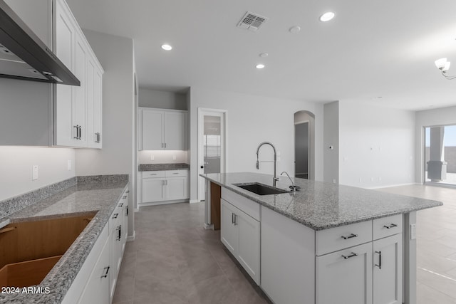 kitchen with light stone countertops, white cabinetry, a kitchen island with sink, light tile floors, and wall chimney range hood