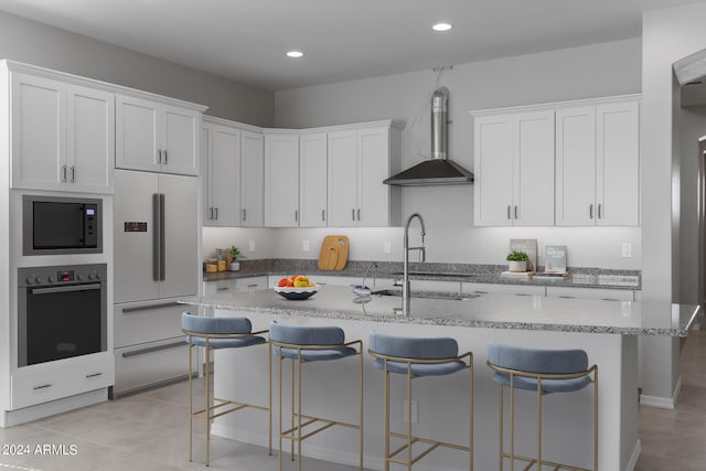 kitchen featuring white cabinetry, built in appliances, wall chimney exhaust hood, and an island with sink