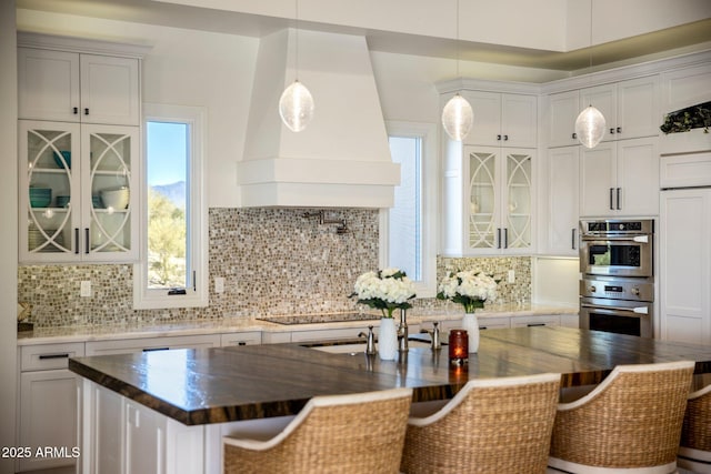 kitchen featuring a kitchen island with sink, white cabinetry, custom range hood, a kitchen bar, and decorative backsplash