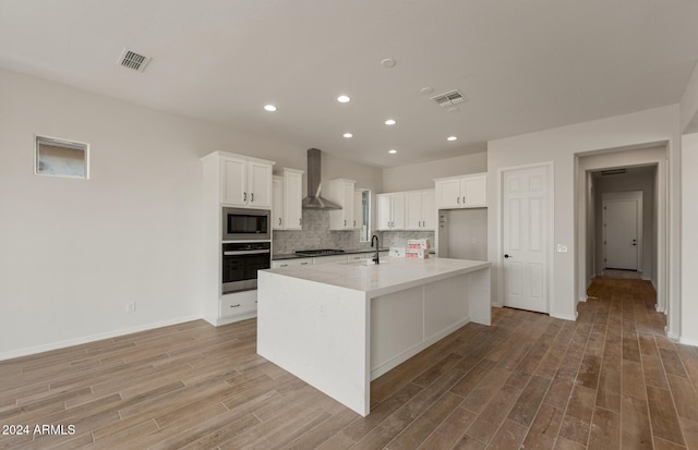 kitchen with stainless steel oven, a center island with sink, white cabinets, wall chimney range hood, and built in microwave