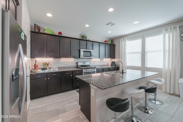 kitchen with a center island with sink, a kitchen breakfast bar, sink, dark stone countertops, and stainless steel appliances