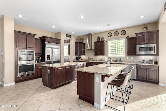 kitchen featuring a center island, wall chimney exhaust hood, sink, built in appliances, and a breakfast bar