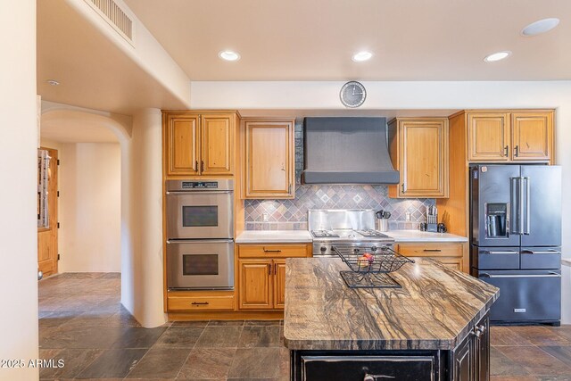 8606 E Arroyo Seco Rd, Scottsdale, AZ 85266 - kitchen featuring stainless steel appliances, wall chimney range hood, arched walkways, backsplash, and dark stone finish floors