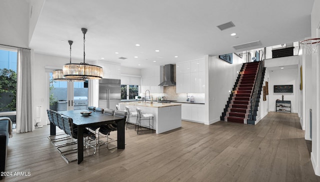 kitchen with white cabinetry, an island with sink, built in refrigerator, wall chimney exhaust hood, and decorative light fixtures