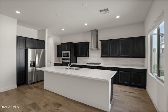 kitchen with an island with sink, sink, wall chimney exhaust hood, stainless steel appliances, and light wood-type flooring