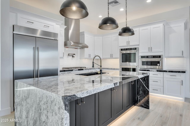 kitchen featuring visible vents, a sink, stainless steel appliances, white cabinetry, and wall chimney range hood