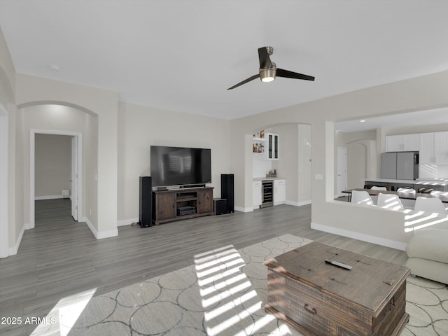 living area featuring arched walkways, beverage cooler, a ceiling fan, baseboards, and light wood-style floors