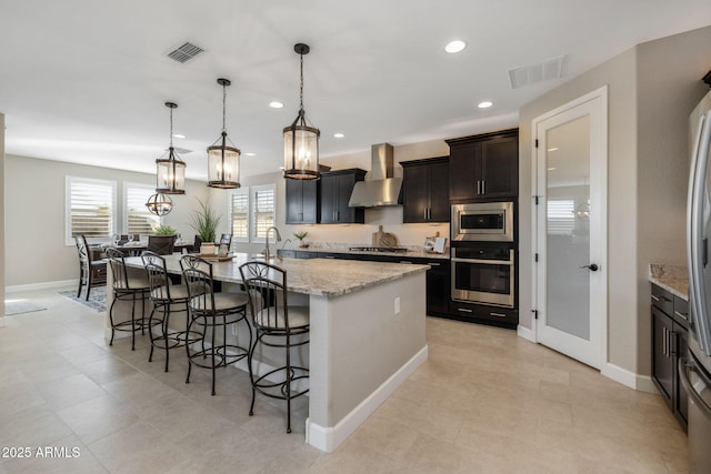 kitchen with stainless steel appliances, wall chimney range hood, decorative light fixtures, and a center island with sink