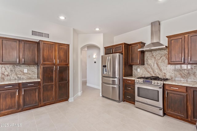 kitchen featuring appliances with stainless steel finishes, decorative backsplash, light stone counters, dark brown cabinets, and wall chimney exhaust hood