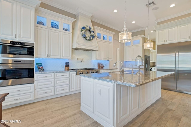 kitchen with appliances with stainless steel finishes, backsplash, white cabinets, a center island with sink, and custom exhaust hood