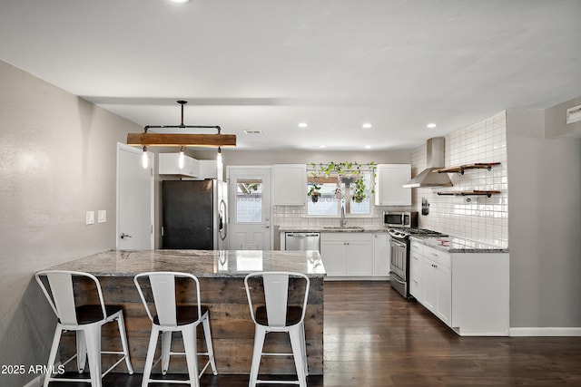 kitchen featuring appliances with stainless steel finishes, a sink, a peninsula, decorative backsplash, and wall chimney range hood