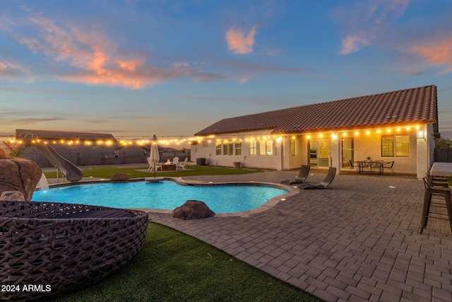 pool at dusk with a patio area, a yard, and a water slide