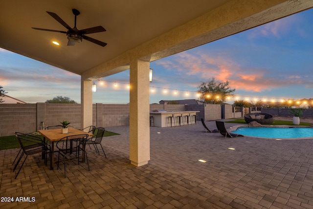 patio terrace at dusk featuring a water view, ceiling fan, a fenced in pool, area for grilling, and an outdoor bar