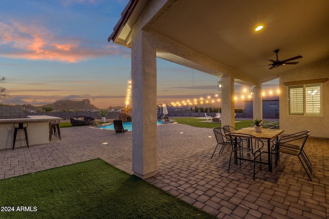 patio terrace at dusk with a lawn, a mountain view, ceiling fan, and exterior bar