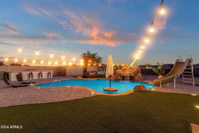 pool at dusk featuring a water slide, a lawn, pool water feature, and a patio area