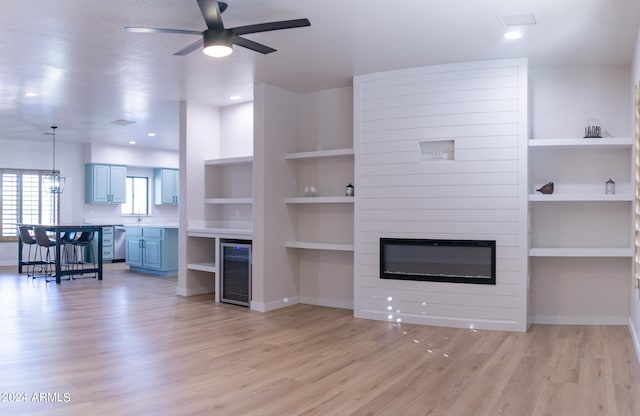 unfurnished living room featuring beverage cooler, built in shelves, light hardwood / wood-style flooring, a fireplace, and ceiling fan with notable chandelier