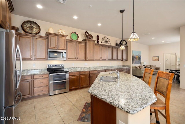 kitchen with a center island with sink, stainless steel appliances, brown cabinetry, a sink, and light stone countertops