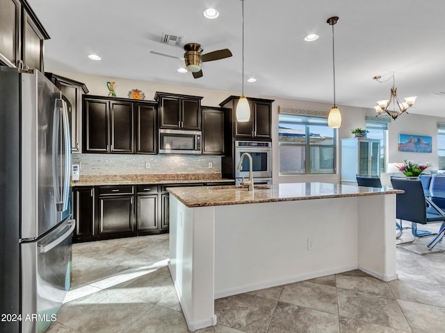 kitchen featuring pendant lighting, a kitchen island with sink, ceiling fan with notable chandelier, appliances with stainless steel finishes, and light stone counters