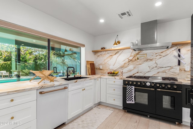 kitchen featuring white dishwasher, range with two ovens, white cabinets, sink, and wall chimney range hood