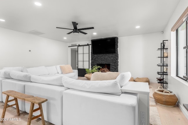 living room featuring a stone fireplace, ceiling fan, a barn door, and light hardwood / wood-style floors