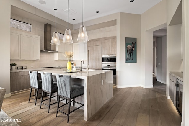 kitchen featuring light brown cabinets, wall chimney exhaust hood, an island with sink, pendant lighting, and wood-type flooring