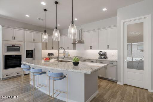 kitchen with wall chimney range hood, hanging light fixtures, light hardwood / wood-style flooring, a kitchen island with sink, and sink