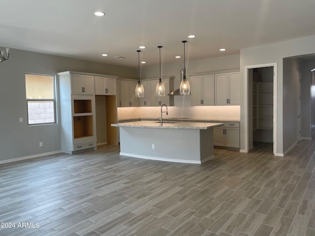 kitchen featuring a center island with sink, light hardwood / wood-style flooring, light stone counters, decorative light fixtures, and backsplash