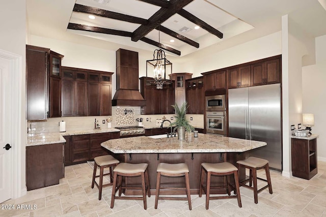 kitchen with custom exhaust hood, beamed ceiling, an island with sink, decorative light fixtures, and built in appliances