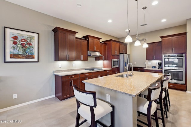 kitchen with sink, hanging light fixtures, light stone counters, an island with sink, and appliances with stainless steel finishes