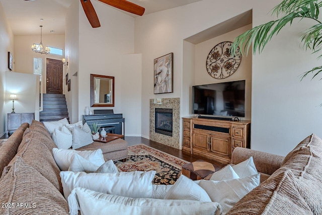 living area featuring a glass covered fireplace, stairway, wood finished floors, high vaulted ceiling, and ceiling fan with notable chandelier