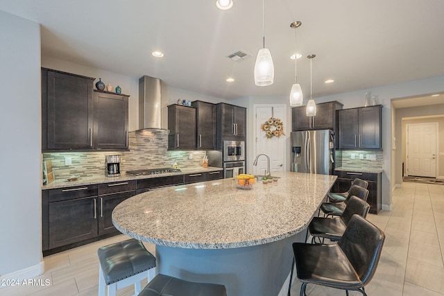 kitchen with a kitchen bar, a kitchen island with sink, wall chimney exhaust hood, and appliances with stainless steel finishes