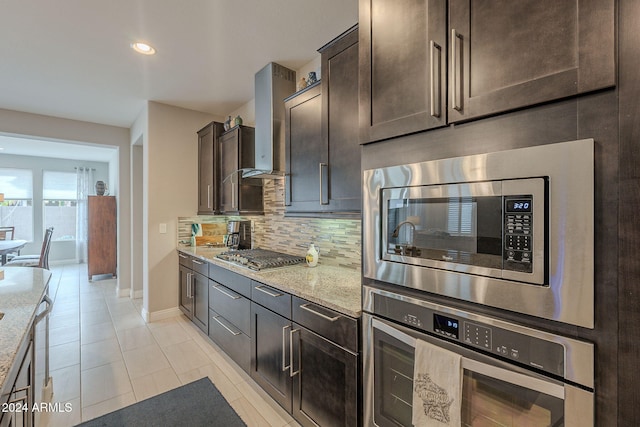 kitchen featuring light stone countertops, appliances with stainless steel finishes, dark brown cabinets, and wall chimney range hood