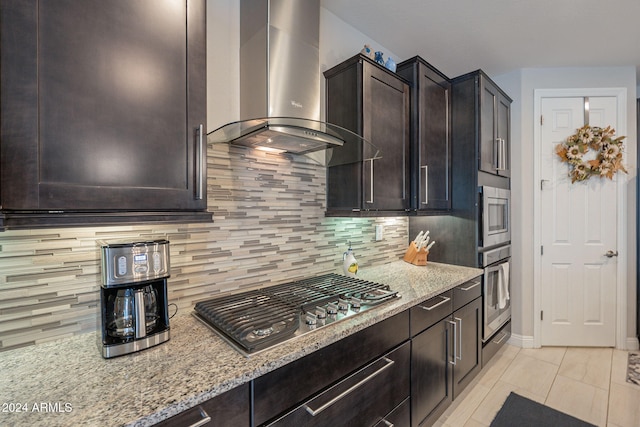 kitchen featuring dark brown cabinetry, wall chimney range hood, tasteful backsplash, light stone counters, and appliances with stainless steel finishes