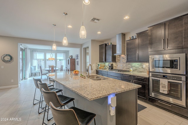 kitchen with sink, an island with sink, decorative light fixtures, light stone counters, and stainless steel appliances