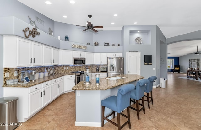 kitchen with white cabinetry, stainless steel appliances, dark stone countertops, ceiling fan, and a center island with sink