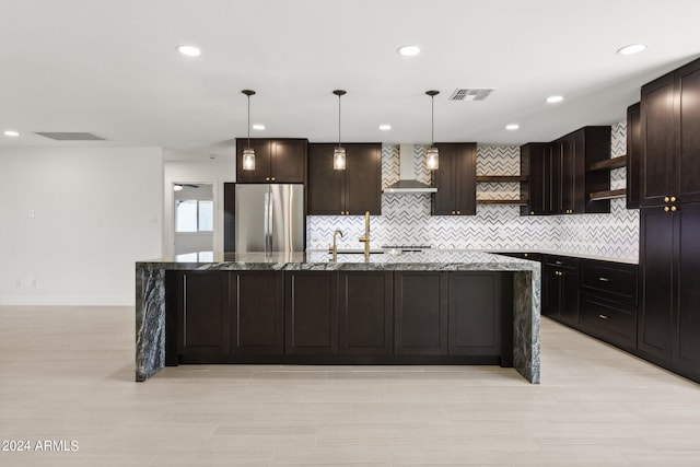 kitchen with a center island with sink, decorative light fixtures, wall chimney range hood, and stainless steel fridge