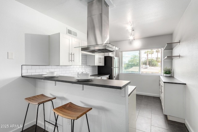 kitchen with dark countertops, visible vents, backsplash, island exhaust hood, and freestanding refrigerator