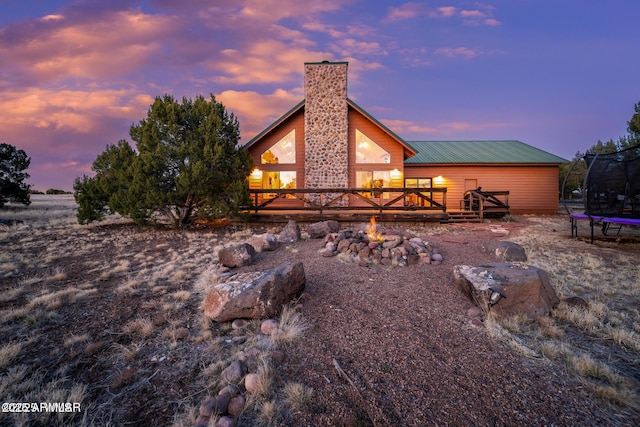 back of house at dusk with a trampoline, a chimney, a fire pit, a wooden deck, and metal roof
