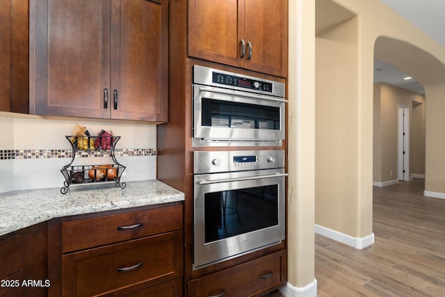 kitchen featuring light stone countertops, arched walkways, stainless steel double oven, light wood finished floors, and baseboards
