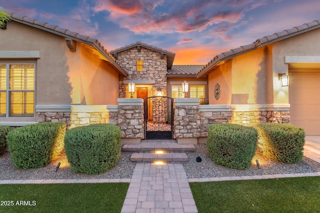 exterior entry at dusk with a tiled roof, stucco siding, stone siding, a gate, and a garage