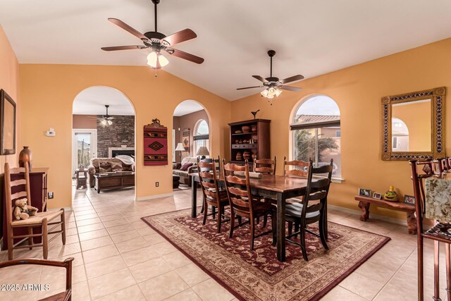 dining area featuring a ceiling fan, arched walkways, a large fireplace, vaulted ceiling, and light tile patterned flooring